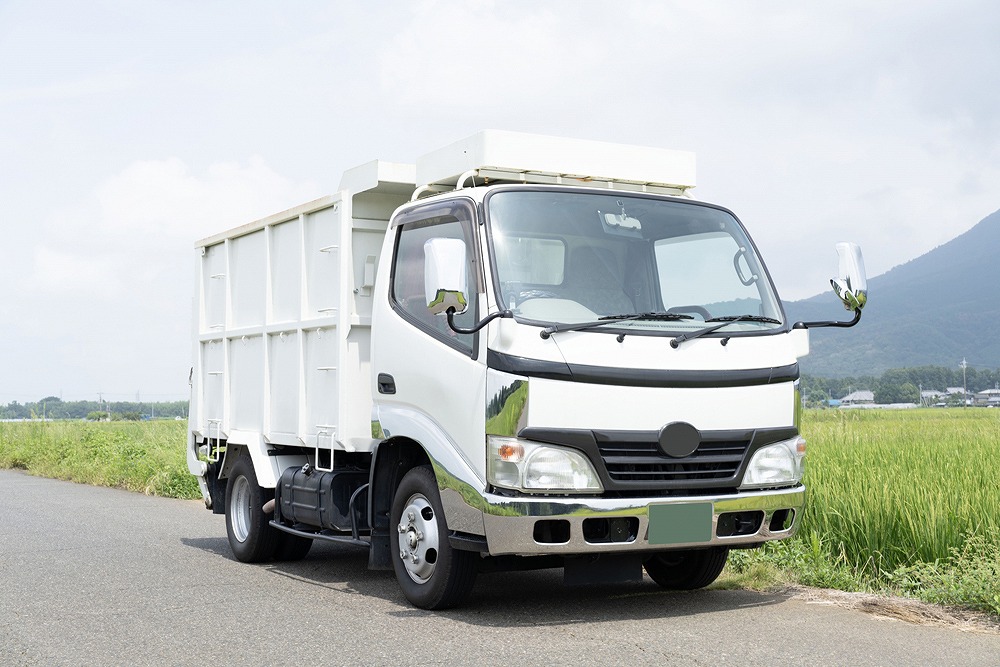 田園地帯の道路を走る白い大型トラック（産業廃棄物や粗大ごみ収集車イメージ）